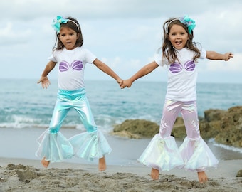 May include: Two girls wearing mermaid costumes, one in blue and one in pink, jump in the air on a sandy beach. They are both smiling and holding hands.