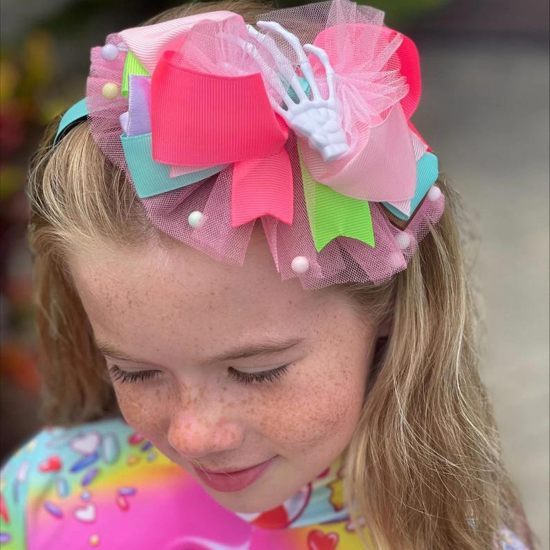 May include: A hair accessory featuring a large, multi-layered bow made of neon pink, lime green, and light blue grosgrain ribbon. The bow is accented with layers of pink tulle, small white pom-poms, and a white plastic skeleton hand charm.
