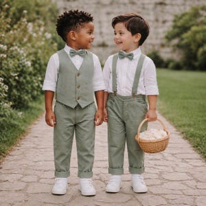 May include: Two young boys in matching sage green outfits, including vests, pants, and bow ties. One boy wears suspenders. Both wear white shirts and sneakers. One carries a small woven basket filled with white flowers. The boys are standing on a stone path.