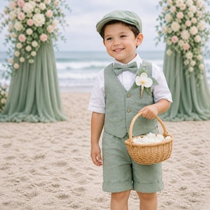 May include: A sage green vest, matching shorts, and a flat cap worn with a white short-sleeved button-down shirt and a matching bow tie. A white orchid boutonniere is pinned to the vest, and a small woven wicker basket holds white flower petals.