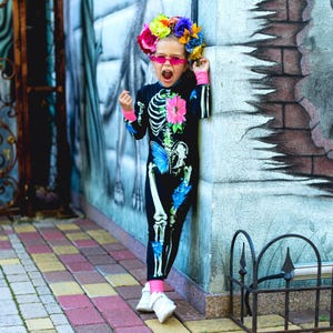 May include: A black long-sleeved jumpsuit featuring a white skeleton print with a pink flower on the chest and a blue butterfly on the hip. The outfit includes pink wrist cuffs, a floral headpiece with colorful roses, and pink flame-shaped sunglasses.