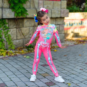 May include: A bright pink long-sleeve jumpsuit featuring a graphic print of a skeleton ribcage filled with colorful sweets, including a heart-shaped candy and a donut at the waist, paired with white sneakers and a large multi-colored hair bow.