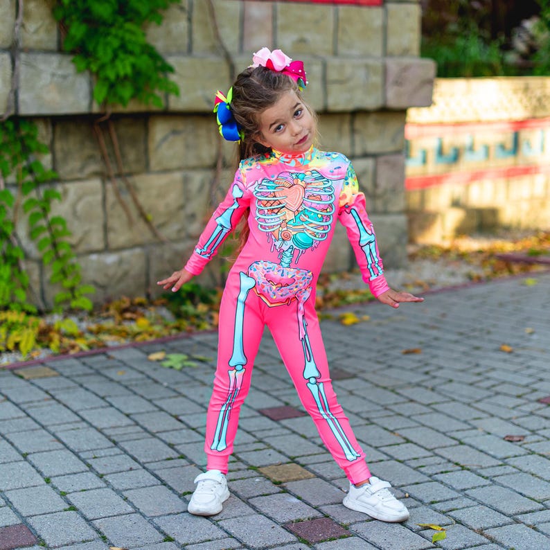 May include: A bright pink long-sleeve jumpsuit featuring a graphic print of a skeleton ribcage filled with colorful sweets, including a heart-shaped candy and a donut at the waist, paired with white sneakers and a large multi-colored hair bow.