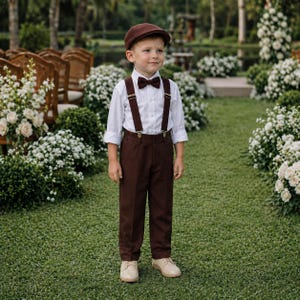 May include: A young person in a formal outfit, including a white button-down shirt, brown pants, suspenders, and a bow tie. The ensemble is completed with a brown cap and beige shoes. The outfit is suitable for a special occasion.