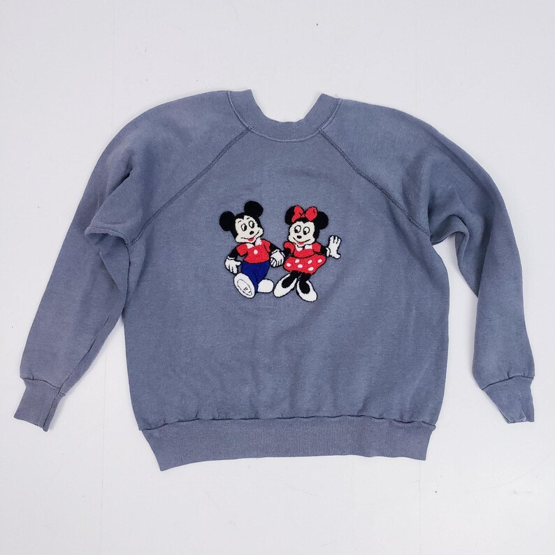 Puede incluir: Sudadera azul gris&aacute;ceo con un bordado de Mickey y Minnie Mouse. Mickey lleva una camisa roja y pantalones azules, Minnie un vestido de lunares rojos. La sudadera tiene mangas largas y cuello redondo.