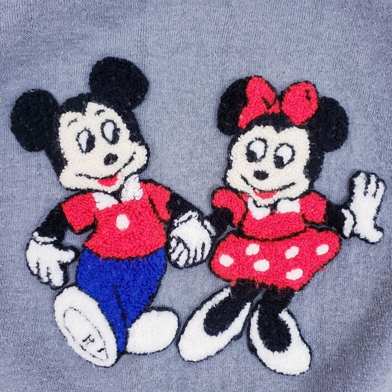 Puede incluir: Personajes bordados de Mickey Mouse y Minnie Mouse sobre un fondo gris. Mickey lleva una camisa roja, pantalones azules y guantes blancos. Minnie lleva un vestido rojo con lunares blancos y un lazo rojo. Los personajes se dan la mano.