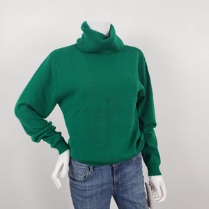 May include: A solid emerald green long-sleeve sweater featuring a high turtleneck collar and ribbed cuffs at the wrists and hem. The sweater is displayed on a mannequin torso paired with blue denim jeans, showcasing a simple and classic design.