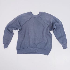 Puede incluir: Una sudadera azul descolorida con mangas largas y cuello redondo. Las mangas ragl&aacute;n se extienden desde el escote hasta la axila. Los pu&ntilde;os y el dobladillo son acanalados. La tela tiene un aspecto ligeramente desgastado.