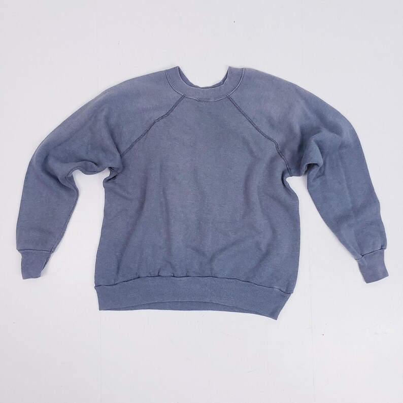 Puede incluir: Una sudadera azul descolorida con mangas largas y cuello redondo. Las mangas ragl&aacute;n se extienden desde el escote hasta la axila. Los pu&ntilde;os y el dobladillo son acanalados. La tela tiene un aspecto ligeramente desgastado.