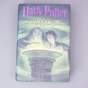 Puede incluir: Un libro de tapa dura titulado Harry Potter and the Half-Blood Prince de J.K. Rowling. La portada presenta una ilustración en verde y negro de un hombre mayor con barba larga y una persona joven con gafas sosteniendo una varita sobre un cuenco.