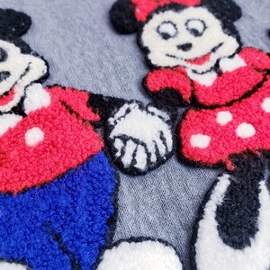 Puede incluir: Primer plano de una tela gris con personajes de dibujos animados bordados de Mickey Mouse y Minnie Mouse. Mickey viste de rojo y azul, Minnie de rojo con lunares blancos. Los personajes se dan la mano.