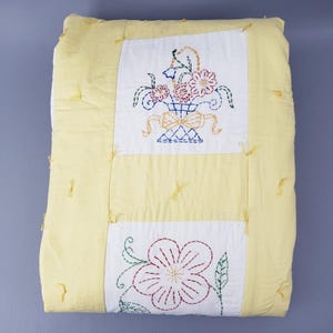 May include: A rectangular quilt featuring a pale yellow border and two white central panels. The top panel displays an embroidered basket of flowers in red, blue, and yellow. The bottom panel shows a large embroidered flower with green leaves.
