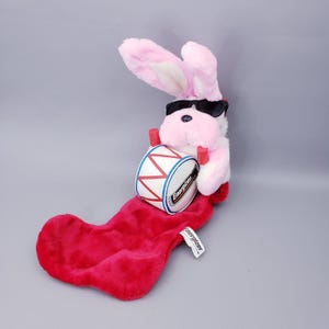 May include: A plush white rabbit with long pink-lined ears wearing black sunglasses. The rabbit holds a white drum with red trim and the Energizer logo, attached to a soft red stocking featuring a small white Energizer brand tag.