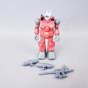 May include: A red and gray robot action figure with the number 108 printed on its chest and feet. The robot has a white head with two antennae and gray shoulder pads. Three gray weapon accessories are included.