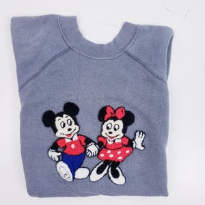 Puede incluir: Una sudadera gris con im&aacute;genes bordadas de Mickey Mouse y Minnie Mouse. Mickey lleva una camisa roja, pantalones azules y guantes blancos. Minnie lleva un vestido rojo con lunares blancos y un lazo rojo. La sudadera est&aacute; doblada.