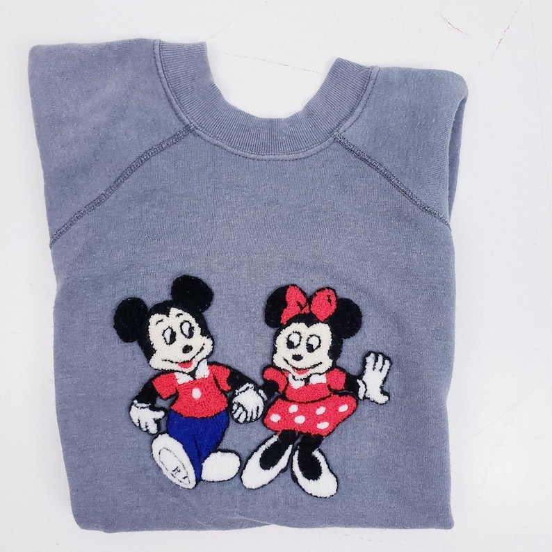 Puede incluir: Una sudadera gris con im&aacute;genes bordadas de Mickey Mouse y Minnie Mouse. Mickey lleva una camisa roja, pantalones azules y guantes blancos. Minnie lleva un vestido rojo con lunares blancos y un lazo rojo. La sudadera est&aacute; doblada.