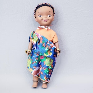 May include: A vintage doll with dark skin, wearing an orange top and a blue patterned shawl. The doll has a beaded headband, necklace, and bracelets. Silver rings adorn the doll's ankles. The doll's clothing has a colorful floral design.