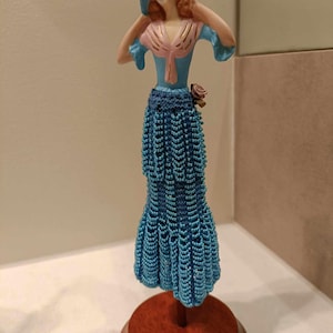 May include: A porcelain doll figurine wearing a blue and pink dress with a beaded skirt and a blue hat with flowers. The doll is standing on a wooden base.