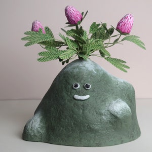 May include: A green, rock-shaped planter with a smiling face and black eyes. The planter has a small, white mouth and is topped with a green plant with pink flowers.