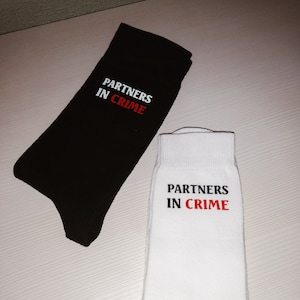Op de afbeelding: Een paar sokken, een zwarte en een witte, met de tekst "Partners in Crime" in rode en witte letters.
