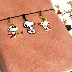 May include: A collection of Snoopy-themed charms with gold-colored clasps. The charms feature Snoopy in various poses, including playing a guitar and wearing a crown. They are displayed on a pink leather-bound notebook.
