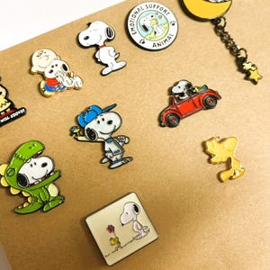 May include: A collection of Snoopy character pins in various poses and designs. Includes Snoopy in a red car, a dinosaur costume, and a baseball cap. Other pins feature Snoopy with Woodstock, and the text "BE YOU! THE WORLD WILL ADJUST".