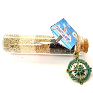 Peut inclure: Une petite bouteille en verre remplie de sable en couches provenant des îles Canaries, avec un bouchon en liège et un charme de boussole attaché.