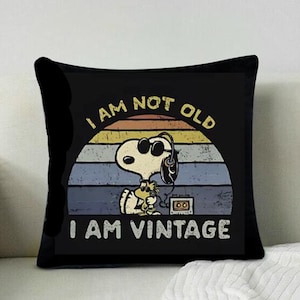 Puede incluir: Cojín cuadrado negro con Snoopy con gafas de sol y auriculares, y el texto "I AM NOT OLD I AM VINTAGE". El diseño incluye una puesta de sol retro y una ilustración de casete.