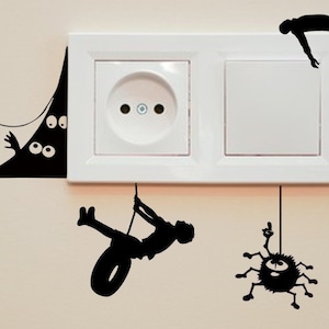 Puede incluir: Una toma de corriente eléctrica blanca y un interruptor de luz con calcomanías de pared en silueta negra de un monstruo que asoma por detrás de una pared, una araña colgando del techo y una persona balanceándose en un columpio de neumático.