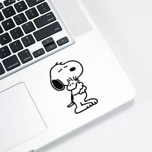 Op de afbeelding: Een zwart-witte Snoopy-sticker is bevestigd op een laptop. De sticker toont Snoopy in een contemplatieve pose, met gesloten ogen en gekruiste armen. De laptop is wit met een zwart toetsenbord.