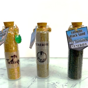 May include: Three small glass bottles filled with different coloured sand from Tenerife, Spain. The bottles are labelled with the names of the beaches and the type of sand. The labels are in Spanish and English.