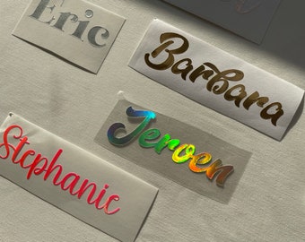 May include: Close-up of several name decals. The names "Eric" in silver, "Barbara" in gold, "Stephanie" in red, and "Jeroen" in iridescent colors are displayed on individual adhesive labels. The background is a light gray surface.