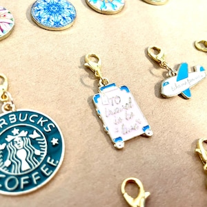 Puede incluir: Una colección de abalorios de esmalte de colores con cierres dorados. Los abalorios incluyen un logotipo de Starbucks, una maleta con el texto "Viajar es vivir", un avión con el texto "Llévame lejos", y algunos otros diseños abstractos.