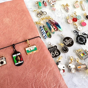 Puede incluir: Una colección de abalorios coloridos y únicos para una agenda o un cuaderno. Los abalorios incluyen una cinta de casete, un logotipo de Bob Marley, un cartel de la Ruta 66, una brújula, una televisión, una bombilla y un logotipo de los Beatles.