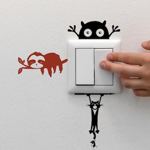 May include: A white light switch with a black and red sloth and cat wall decals. A hand is reaching to turn on the light switch. The sloth is resting on a branch, and the cat is hanging from the switch.