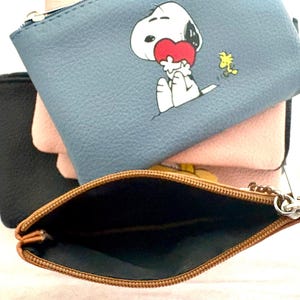 Könnte beinhalten: Ein Stapel kleiner Reißverschlusstaschen in verschiedenen Farben. Die obere Tasche ist blau mit einer Snoopy-Grafik, die ein Herz hält. Andere Taschen sind rosa und schwarz. Die offene Tasche unten hat einen braunen Reißverschluss und Innenraum.