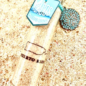 Peut inclure: Une bouteille en verre transparente remplie de sable, étiquetée "Icaos Puerto Rico". Une étiquette bleue et un charme décoratif en forme de dollar des sables sont attachés. La bouteille repose sur une surface sablonneuse.