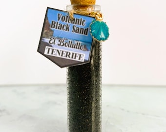 Puede incluir: Una pequeña botella de vidrio llena de arena negra, sellada con un corcho y un dije turquesa. Una etiqueta dice "Volcanic Black Sand El Bollullo Tenerife."
