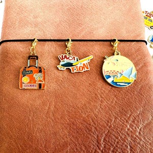 Puede incluir: Tres colgantes con temática de viaje cuelgan de una banda elástica negra. Los colgantes incluyen una maleta naranja con las palabras "Travel" y "Hello", un avión con "Vacation" y un colgante redondo con una escena de playa.