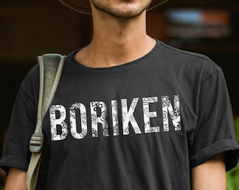 Puede incluir: Una persona con una camiseta negra con la palabra "BORIKEN" impresa en blanco. La camiseta tiene un diseño estampado en blanco. La persona lleva un sombrero marrón y mira hacia arriba.