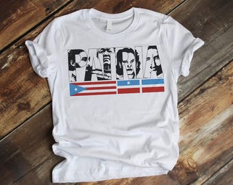 May include: White t-shirt with a black and white graphic of the word "PATRIA" with images of people's faces incorporated into the letters. The graphic also includes three red, white, and blue stripes with a blue star in each stripe.