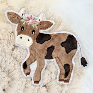 Könnte beinhalten: Eine braune und weiße Kuh mit einer gestickten Blumenkrone auf dem Kopf. Die Kuh steht auf einem weißen Hintergrund.
