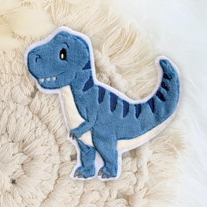 Könnte beinhalten: Ein blaues Plüschtier, das einen Tyrannosaurus Rex darstellt, mit dunkelblauen Streifen und weißen Akzenten. Der Dinosaurier hat einen freundlichen Ausdruck und ist aus weichem Stoff gefertigt. Ein niedliches, kuscheliges Spielzeug für Kinder.