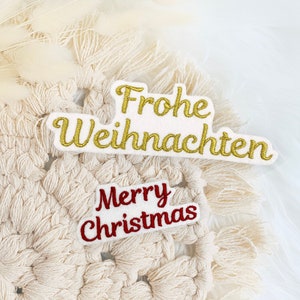 Könnte beinhalten: Weißer gehäkelter Wandbehang mit zwei gestickten Patches. Der obere Patch sagt "Frohe Weihnachten" in goldener Farbe. Der untere Patch sagt "Merry Christmas" in roter Farbe.