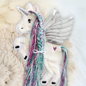 Könnte beinhalten: Weißes Einhorn-Applikation mit silbernem Horn und Flügeln. Das Einhorn hat eine geflochtene Mähne aus rosa, türkis und silbernen Strähnen und einen passenden Schweif. Ein kleines rosa Herz befindet sich am Hinterteil des Einhorns.