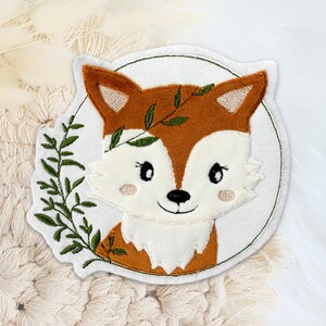 Könnte beinhalten: Ein weißer Filz-Patch mit einem gestickten braunen Fuchs. Der Fuchs hat einen grünen Zweig mit Blättern um den Kopf. Der Patch ist von einem grünen Kreis umgeben.