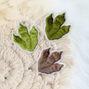 Könnte beinhalten: Drei Dinosaurier-Fußabdrücke aus Filz in Grün und Braun. Die Fußabdrücke befinden sich auf einer cremefarbenen gehäkelten Oberfläche.