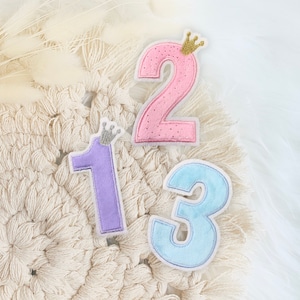 Könnte beinhalten: Drei Filz-Zahlen, 1, 2 und 3, mit einer Krone auf jeder Zahl. Die Zahlen sind in verschiedenen Farben: lila, rosa und blau.