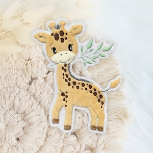Könnte beinhalten: Eine gestickte Giraffen-Applikation in Braun und Weiß mit braunen Flecken und einem grünen Blatt im Maul. Die Applikation befindet sich auf einem weißen Hintergrund.