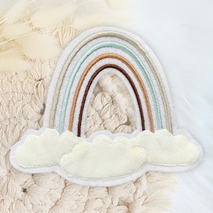Könnte beinhalten: Ein regenbogenfarbener Filz-Patch mit weißem Hintergrund und zwei weißen Wolken. Der Regenbogen hat fünf Streifen in den Farben Braun, Orange, Grün, Hellblau und Weiß.